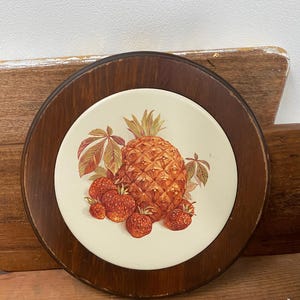 May include: Round wooden trivet with a cream-colored ceramic center. The ceramic features a detailed illustration of a pineapple and several strawberries in shades of orange, red, and green. The trivet has a dark brown wooden frame.