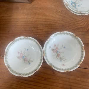 May include: Two small white porcelain bowls with a green and gold floral pattern. The bowls have a scalloped edge and are decorated with pink and blue flowers.