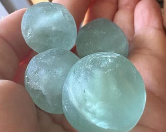 Op de afbeelding: Vier afgeronde, doorschijnende glazen bollen in een bleke zeegroene kleur met een gladde, verweerde textuur. Elke bol vertoont subtiele interne variaties en een zacht, mat uiterlijk dat doet denken aan glas dat door de zee is gepolijst.