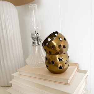 May include: Two stacked, gold-colored metal candle holders with star cutouts. A clear glass oil lamp and a white ceramic lamp are also visible. The items are arranged on a stack of books with cream-colored pages, creating a cozy, vintage aesthetic.