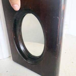 May include: A rectangular dark brown wooden mirror frame with a smooth, rounded finish and a central oval-shaped glass mirror. The wood displays visible grain and slight wear along the edges, featuring a deep, rich tone throughout the structure.
