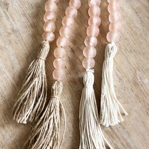 May include: Two strands of pink glass beads with tassels. One tassel is made of natural jute and the other is made of white cotton.
