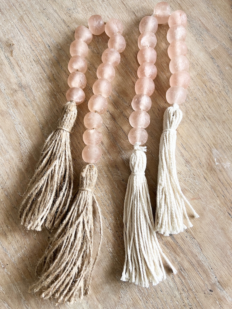 May include: Two strands of pink glass beads with tassels. One tassel is made of natural jute and the other is made of white cotton.