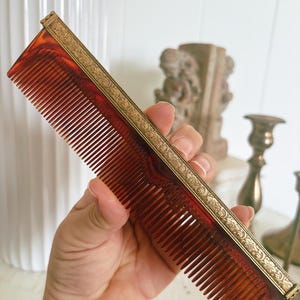 May include: A rectangular hair comb featuring a translucent amber-colored body with fine teeth and a decorative metallic top edge. The metal spine displays an intricate embossed floral pattern that runs along the entire length of the comb.