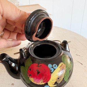 May include: A black ceramic teapot with a lid, featuring hand-painted fruit designs including a red apple, yellow pear, and blue berries. The teapot has a small spout and a handle, and is set on a white surface.