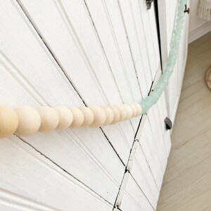 May include: A white wooden door with a beaded garland. The garland is made of light blue and natural wood beads. The beads are strung together on a white string.