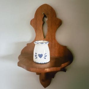 May include: A small white ceramic jar with a blue painted heart and floral pattern sits on a wall-mounted wooden shelf. The shelf features a curved, decorative backboard with a central cutout and a flat surface for holding small objects.