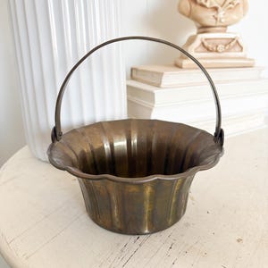 May include: A small, antique brass-colored metal basket with a handle. The basket has a scalloped rim and vertical ribbed detailing. The handle is a dark metal. The basket sits on a distressed, off-white table.