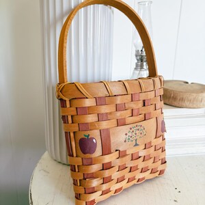 May include: Woven rectangular basket with a handle. The basket is made of light brown and reddish-brown woven strips. It features a painted apple and tree design on the front. The handle is made of a single strip of light brown wood.