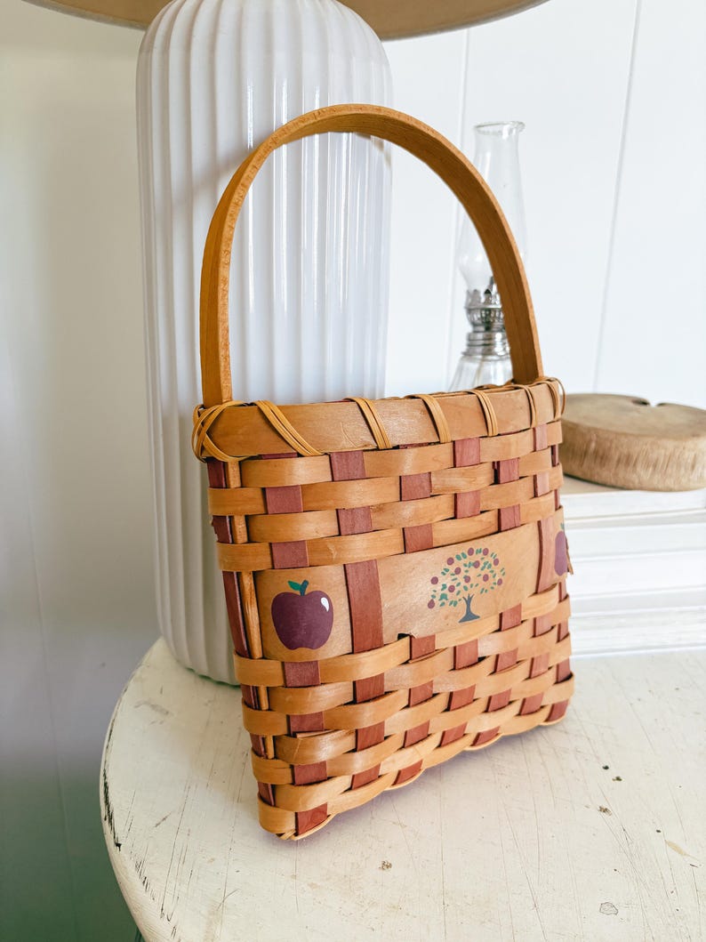 May include: Woven rectangular basket with a handle. The basket is made of light brown and reddish-brown woven strips. It features a painted apple and tree design on the front. The handle is made of a single strip of light brown wood.