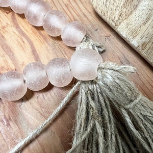 May include: A strand of pale pink glass beads with a natural jute tassel. The beads are strung on a thin twine and the tassel is made of natural jute fibres.