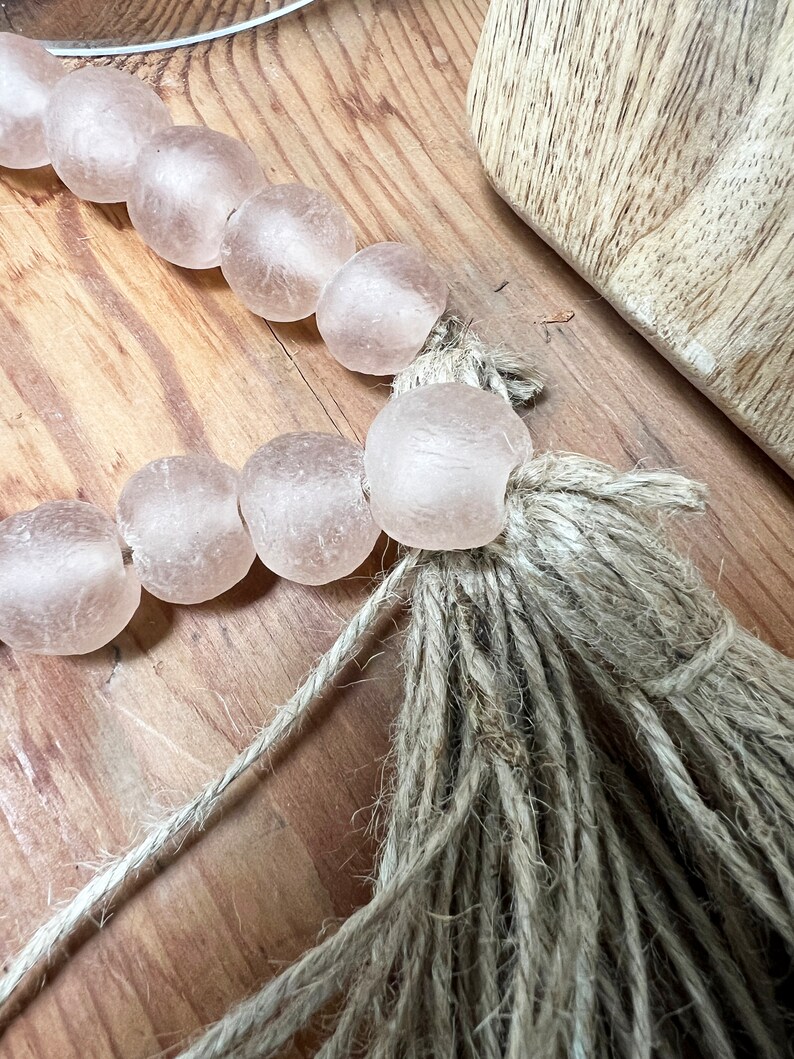 May include: A strand of pale pink glass beads with a natural jute tassel. The beads are strung on a thin twine and the tassel is made of natural jute fibres.