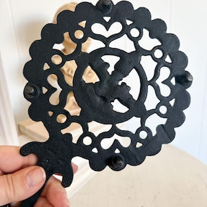 May include: Black cast iron trivet with a decorative, openwork design. The trivet features a circular shape with a scalloped edge and a handle. The design includes hearts and other shapes. The trivet has small feet.