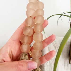 May include: A strand of light pink glass beads with a natural fibre tassel. The beads are round and have a frosted finish.