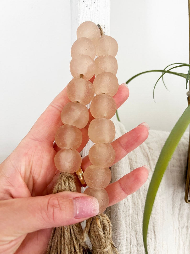 May include: A strand of light pink glass beads with a natural fibre tassel. The beads are round and have a frosted finish.