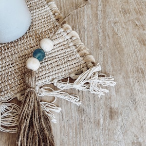 May include: A brown woven placemat with a fringe border and a wooden bead tassel with blue and white beads. The placemat is on a light brown wooden surface.