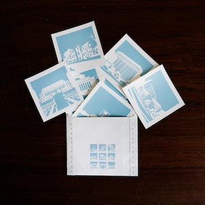 Puede incluir: Un sobre blanco con un borde azul cosido. El sobre est&aacute; lleno de postales azules y blancas con diferentes paisajes urbanos.