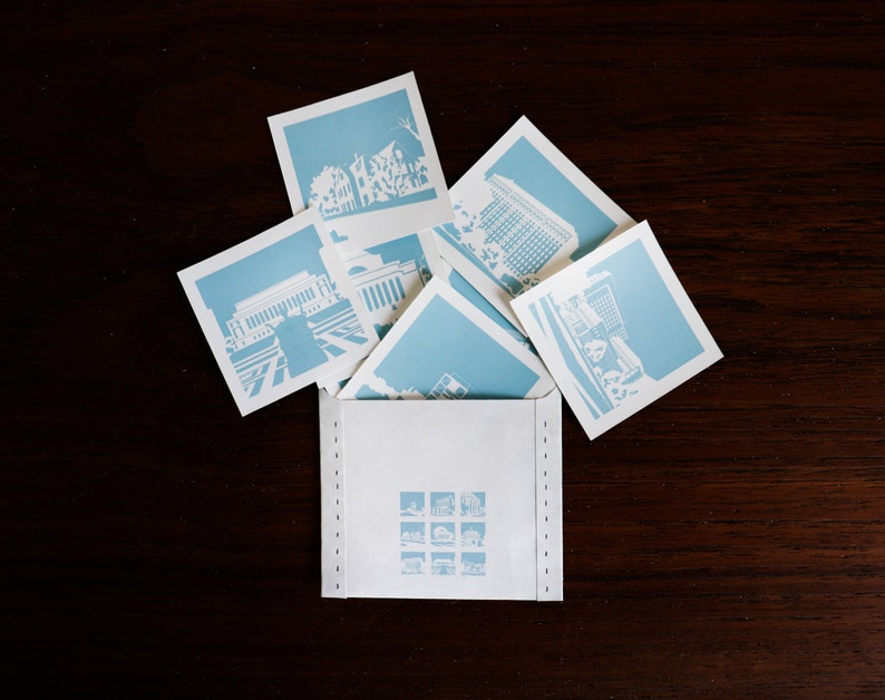 Puede incluir: Un sobre blanco con un borde azul cosido. El sobre est&aacute; lleno de postales azules y blancas con diferentes paisajes urbanos.