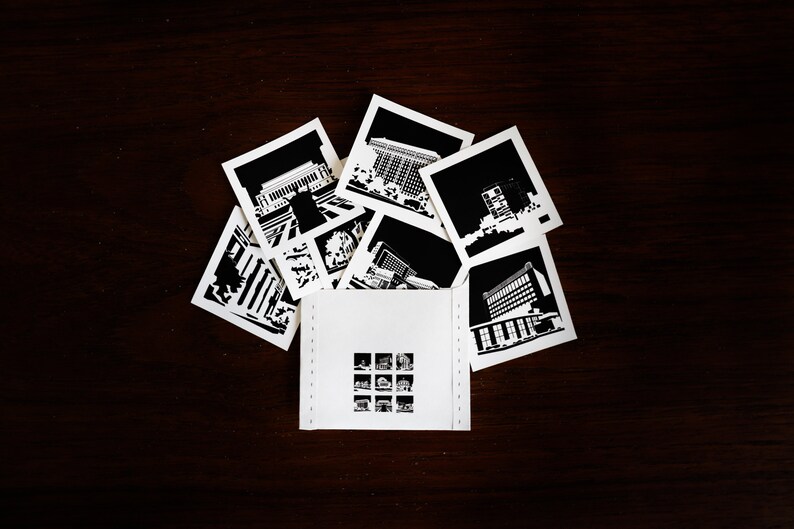 Puede incluir: Un conjunto de fotograf&iacute;as en blanco y negro de edificios, dispuestas en forma de abanico sobre una superficie de madera marr&oacute;n oscuro. Las fotograf&iacute;as est&aacute;n dispuestas alrededor de una tarjeta blanca con una cuadr&iacute;cula de nueve fotograf&iacute;as en blanco y negro m&aacute;s peque&ntilde;as.