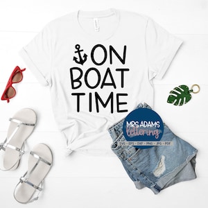 Op de afbeelding: Wit T-shirt met zwarte tekst die "On Boat Time" zegt en een klein ankersymbool. Het shirt ligt op een wit houten oppervlak met een rode zonnebril, een paar zilveren sandalen, een spijkerbroekje en een groen blad.