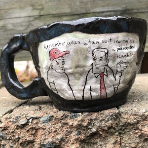 May include: A black ceramic mug with a white and black illustration of two people. The text on the mug reads "Remember when a tax cut counted as a presidential scandal?"