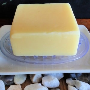 Pode incluir: Uma barra retangular de sabão amarelo claro descansando em um saboneteira de cerâmica branca com um inserto de drenagem de plástico transparente.