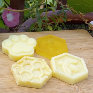 Puede incluir: Cuatro jabones de diferentes formas, cada uno con un diseño diferente. Los jabones son blancos y amarillos y tienen diseños de una huella de pata, un panal y dos abejas. Los jabones están sobre una superficie de madera.