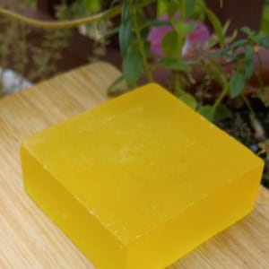 Pode incluir: Uma barra de sabão amarelo brilhante sobre uma superfície de madeira. O sabão é retangular e tem um acabamento liso e brilhante.