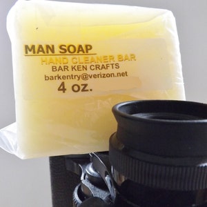 Pode incluir: Uma barra de sabão amarela embrulhada em plástico transparente com o texto "MAN SOAP HAND CLEANER BAR BAR KEN CRAFTS barkentry@verizon.net 4 oz." impresso no rótulo.