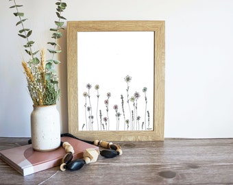 May include: A framed black and white line drawing of a field of flowers with colorful accents. The flowers are drawn in a simple, minimalist style. The frame is a light wood color.