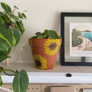 May include: A terracotta flower pot with a hand-painted design of sunflowers. The pot is filled with a green plant with round leaves.
