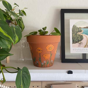 May include: A terracotta flower pot with a hand-painted design of orange poppies and green stems. The pot is sitting on a shelf with a green plant and a framed picture.
