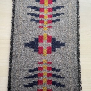 May include: Rectangular gray woven textile with a geometric pattern in red, yellow, and navy blue. The design features a central vertical line with square and arrow-like shapes. The edges are finished with a black fringe.