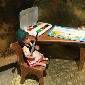 Puede incluir: Una mu&ntilde;eca vestida con un sombrero verde, una camisa blanca y una falda de cuadros rojos y blancos est&aacute; sentada en una silla de madera sosteniendo un pincel. Una mesa de madera con un juego de acuarelas, una paleta de pintura y un dibujo de un paisaje est&aacute; frente a la mu&ntilde;eca.