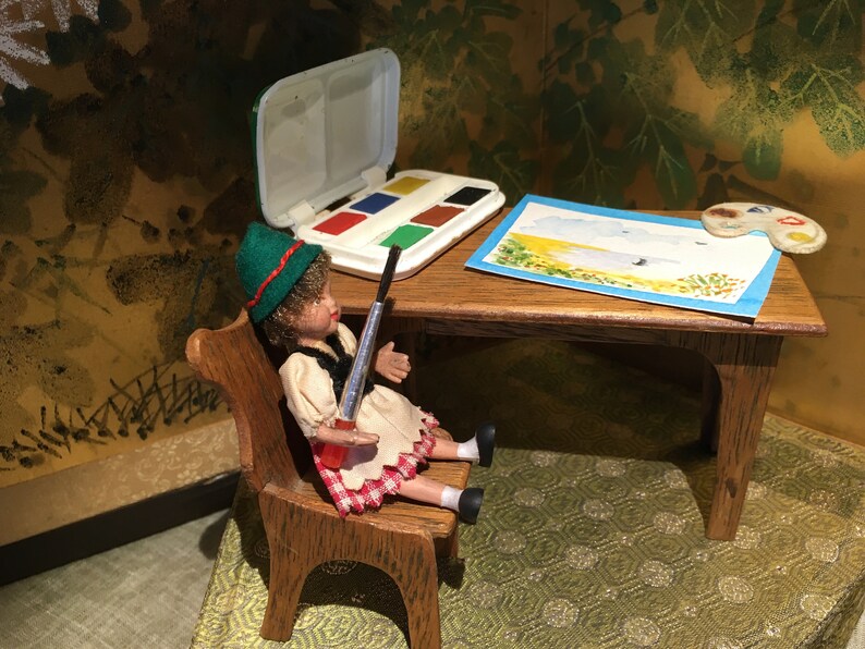 Puede incluir: Una mu&ntilde;eca vestida con un sombrero verde, una camisa blanca y una falda de cuadros rojos y blancos est&aacute; sentada en una silla de madera sosteniendo un pincel. Una mesa de madera con un juego de acuarelas, una paleta de pintura y un dibujo de un paisaje est&aacute; frente a la mu&ntilde;eca.