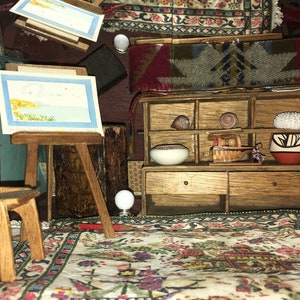 Puede incluir: Una habitaci&oacute;n de casa de mu&ntilde;ecas en miniatura con un caballete de madera, una silla, una peque&ntilde;a pintura, una alfombra, un estante con un taz&oacute;n, una cesta y una maceta, y un peque&ntilde;o globo terr&aacute;queo blanco.