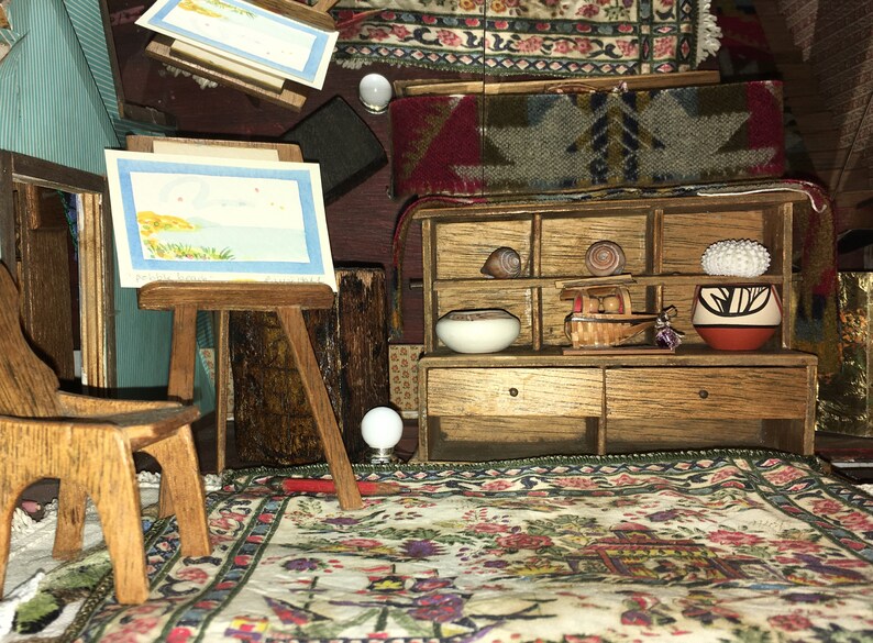 Puede incluir: Una habitaci&oacute;n de casa de mu&ntilde;ecas en miniatura con un caballete de madera, una silla, una peque&ntilde;a pintura, una alfombra, un estante con un taz&oacute;n, una cesta y una maceta, y un peque&ntilde;o globo terr&aacute;queo blanco.