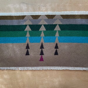 May include: A woven rug with a geometric pattern of triangles and trees. The rug is beige with green, blue, and brown accents. The rug has a fringed edge.