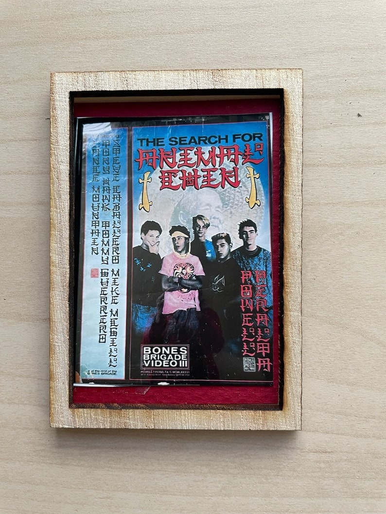Op de afbeelding: Een ingelijste print van een vintage VHS-tapehoes. De hoes toont een groep jonge mannen en de titel "THE SEARCH FOR" in het Engels en Aziatische karakters. De lijst is van licht hout en de passe-partout is rood.