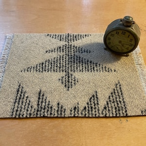 May include: A small, round, silver-colored alarm clock with black numbers and hands. The clock is sitting on a beige rug with a black geometric design.