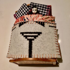 May include: A doll with black hair and a white face is lying in a bed with a black and white checkered pillow and a floral patterned blanket. The bed is made of wood and has a white blanket with a black geometric design.