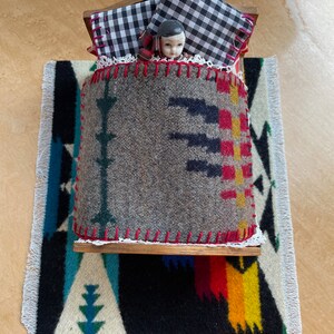 May include: A miniature doll bed with a black and white checkered pillow, a gray blanket with a colorful geometric pattern, and a porcelain doll lying in bed.