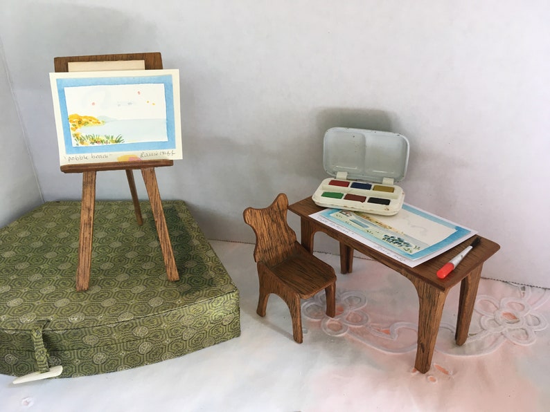 Puede incluir: Un caballete de madera en miniatura con una pintura de acuarela de un paisaje mar&iacute;timo, un peque&ntilde;o escritorio de madera con una silla, un juego de pintura de acuarela y un pincel.