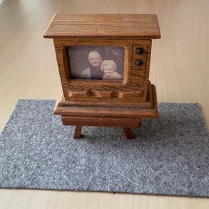 May include: A miniature wooden television with a black and white photo inside. The TV has a vintage design with knobs and a stand. It sits on a gray felt mat, with a light-colored wooden surface in the background.