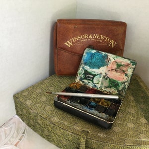 Puede incluir: Un juego de pintura de acuarela vintage Winsor & Newton con una caja de cuero marr&oacute;n. El juego de pintura est&aacute; abierto y muestra una variedad de colores, incluyendo azul, verde y rosa. El juego est&aacute; sobre una superficie con estampado verde y dorado.