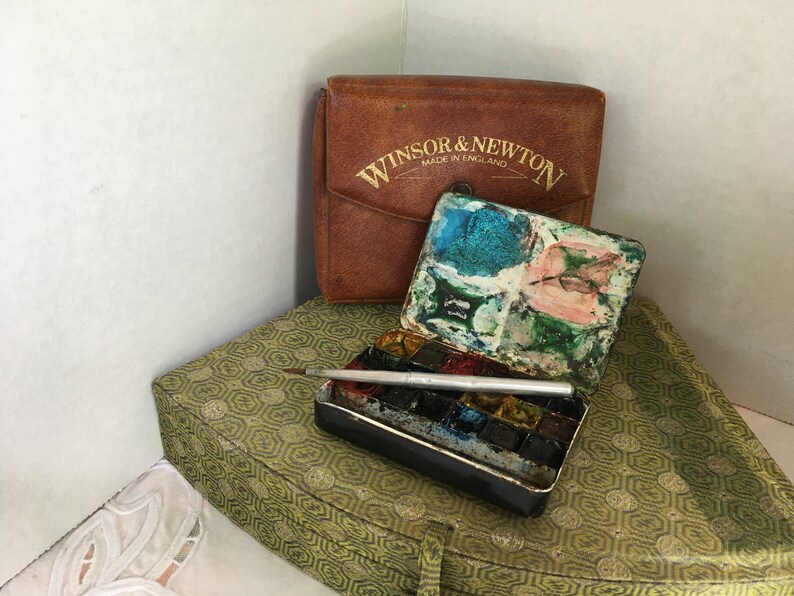 Puede incluir: Un juego de pintura de acuarela vintage Winsor & Newton con una caja de cuero marr&oacute;n. El juego de pintura est&aacute; abierto y muestra una variedad de colores, incluyendo azul, verde y rosa. El juego est&aacute; sobre una superficie con estampado verde y dorado.