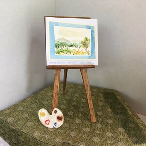 Puede incluir: Un caballete de madera en miniatura con una pintura de acuarela de una escena de paisaje. La pintura est&aacute; titulada "Costa Bon Jour Dreamland" y firmada "Artissana". Una peque&ntilde;a paleta de pintura de madera con pintura amarilla, roja, azul y marr&oacute;n est&aacute; en primer plano.