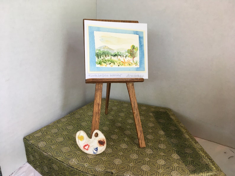 Puede incluir: Un caballete de madera en miniatura con una pintura de acuarela de una escena de paisaje. La pintura est&aacute; titulada "Costa Bon Jour Dreamland" y firmada "Artissana". Una peque&ntilde;a paleta de pintura de madera con pintura amarilla, roja, azul y marr&oacute;n est&aacute; en primer plano.
