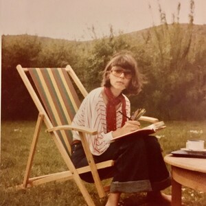 Puede incluir: Una mujer con una camisa a rayas rojas y blancas y gafas de sol marrones se sienta en una silla de playa de madera con un cuaderno y l&aacute;pices. Est&aacute; sentada en un c&eacute;sped verde con una mesa de madera detr&aacute;s de ella.