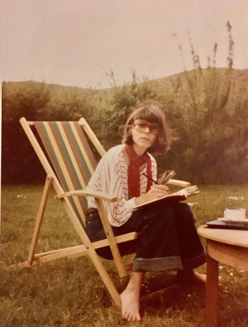 Puede incluir: Una mujer con una camisa a rayas rojas y blancas y gafas de sol marrones se sienta en una silla de playa de madera con un cuaderno y l&aacute;pices. Est&aacute; sentada en un c&eacute;sped verde con una mesa de madera detr&aacute;s de ella.